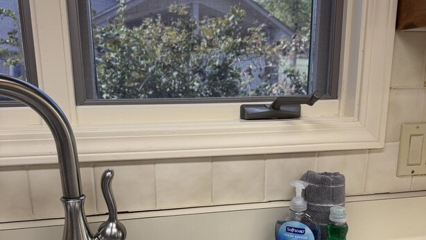 A kitchen sink with a stainless steel faucet, a window with a view of trees and a house, and two bottles of dish soap on the counter. - Birdie House on Balboa (Hot Springs Village)
