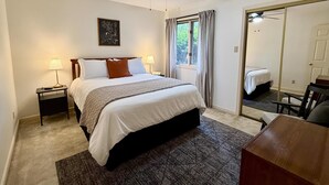 A bedroom with a bed, two bedside tables, a ceiling fan, a window with curtains, a mirror, and a desk with a chair. - Birdie House on Balboa (Hot Springs Village)