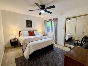 A bedroom with a bed, two bedside tables, a ceiling fan, a window with curtains, a mirror, and a desk with a chair. - Birdie House on Balboa (Hot Springs Village)