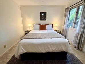 Habitación de hotel con una cama grande, dos mesitas de noche con lámparas, un cuadro enmarcado en la pared y una ventana con cortinas.