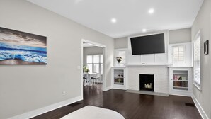 Un salon spacieux doté d’une cheminée, d’un téléviseur à écran plat et d’un coin repas visible à travers une porte ouverte.
