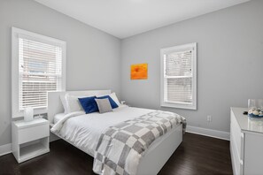 A bedroom with a bed, nightstands, a desk, and two windows with blinds. - Near Monon Trail & Fairgrounds | Meridian Kessler (Indianapolis)