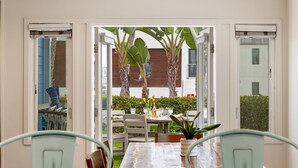 Un espace repas à aire ouverte, avec une table dressée pour deux personnes, entourée de chaises, et une vue sur le jardin extérieur, où l’on aperçoit des bananiers et une haie.