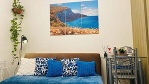 Ein Bett mit blauer Stegdecke, ein an der Wand hängendes Bild von einer Küstenlandschaft, eine hängende Pflanze und ein Schreibtisch mit Stuhl.