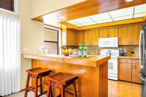 A kitchen with wooden cabinets, a white stove, a microwave, and a refrigerator. There are two wooden stools at the counter. - Kapalua Ridge | Ocean View 3 BR, Sleeps 12 | Car Incl. w/ 6+ Nights | KRV ML-3663 by KBM (Lahaina)