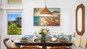 Un coin repas avec une table en bois dressée pour quatre personnes, un tableau représentant une scène de plage au-dessus, et un grand miroir accroché au mur.