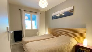 Une chambre avec deux lits, une fenêtre avec des rideaux, un luminaire au plafond, un téléviseur fixé au mur et une table de chevet avec des livres.