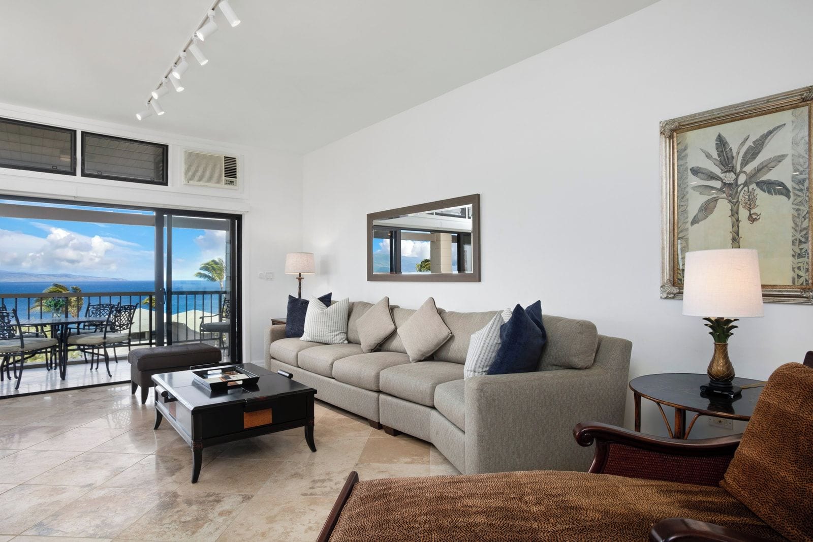 A living room with a sofa, coffee table, and armchairs, a dining area with chairs, a balcony with a view of the ocean, and a painting on the wall.