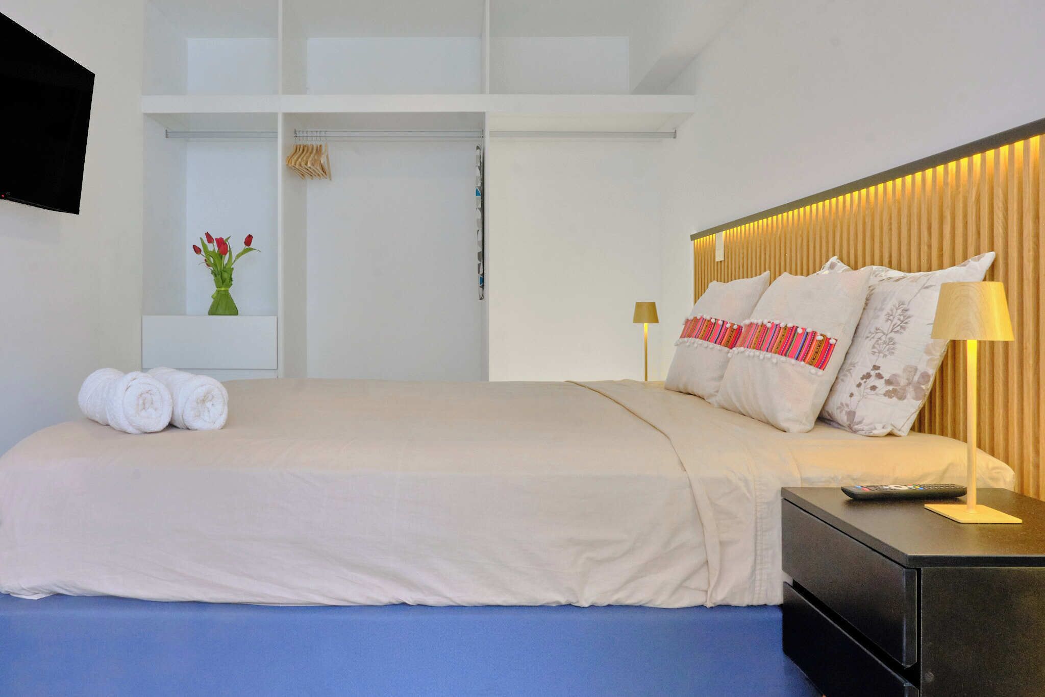 Une chambre d’hôtel moderne équipée d’un lit, d’une table de chevet, d’une télévision et d’une étagère avec des serviettes et un vase.