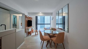 Una cocina moderna con televisor integrado, un comedor con mesa de vidrio y sillas naranjas, y un ventanal con vistas a la ciudad.