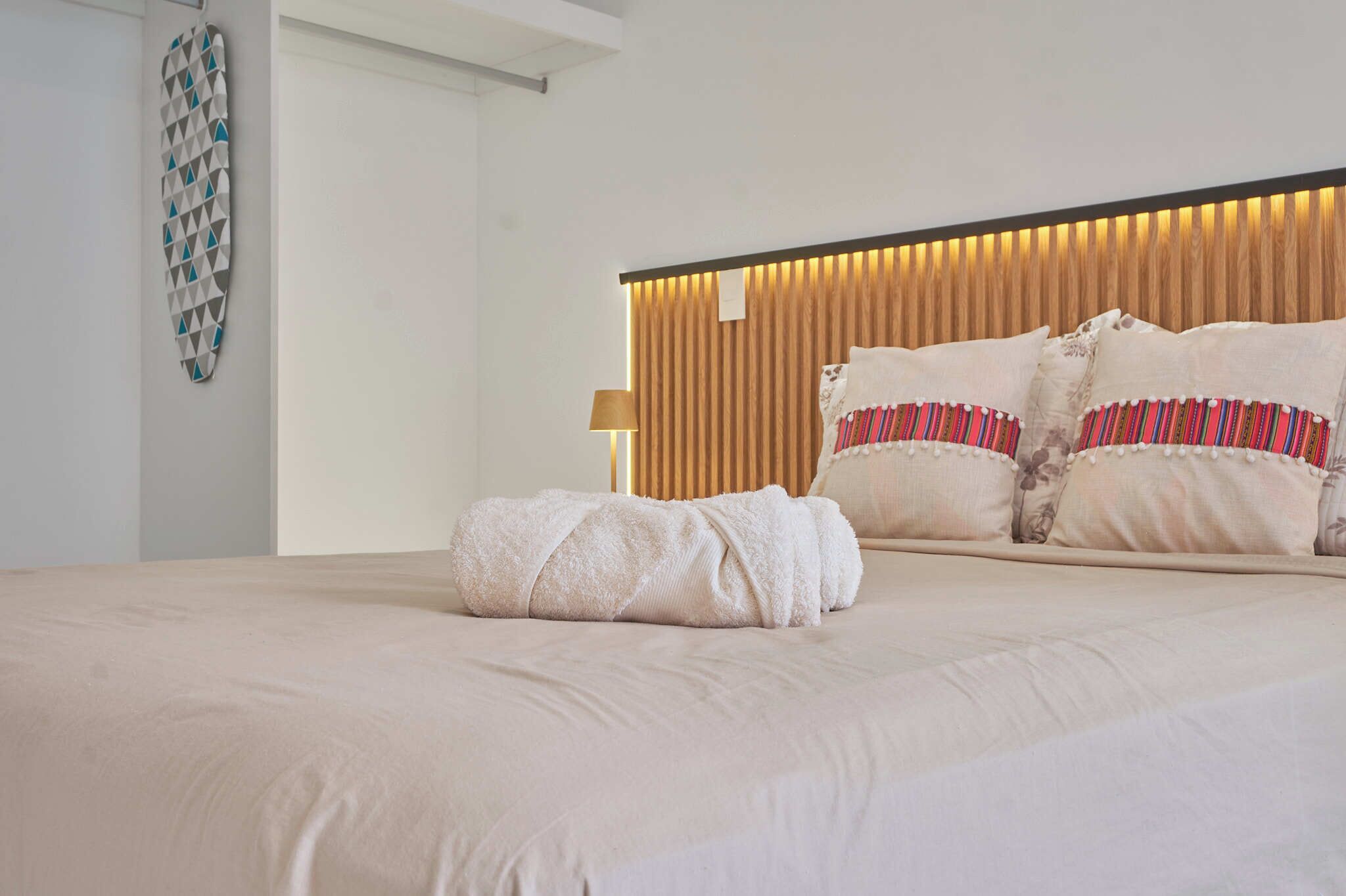 A neatly made bed with a rolled towel, a headboard with vertical wooden slats, and a wall-mounted geometric decoration.