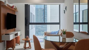 Una sala de estar moderna con un televisor de pantalla plana, una mesa de centro de vidrio, sillas color naranja y un ventanal con vistas a la ciudad.