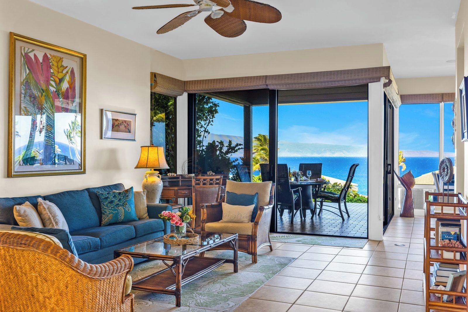 A living room with a sofa, coffee table, and chairs, a ceiling fan, and a view of the ocean through large glass doors.