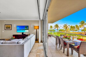 Ein Balkon mit Blick auf Palmen und Meer, eine Sitzecke mit Sofa, ein Fernseher und ein Esstisch für zwei Personen.
