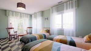 Ein Schlafzimmer mit zwei Betten, einem Schaukelstuhl, einem kleinen Tisch und einem Fenster mit durchsichtigen Vorhängen.