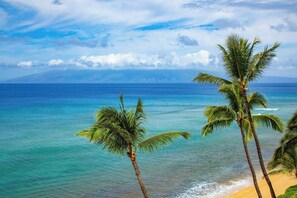 Ein Strand mit klarem türkisblauem Wasser, Palmen und einem fernen Gebirgszug unter einer teils bewölkten Himmel.