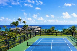 A tennis court with a net, surrounded by a fence, with a view of the ocean and houses in the background. - Kapalua Bay | Ocean View 4 BR, Sleeps 14 | Car Incl. w/ 6+ Nights | KBV ML-3725 by KBM (Lahaina)