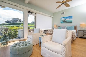 A bedroom with a bed, two chairs, a side table, a lamp, a ceiling fan, and a large window with a view of a building and greenery. - Kapalua Bay | Ocean View 4 BR, Sleeps 14 | Car Incl. w/ 6+ Nights | KBV ML-3725 by KBM (Lahaina)