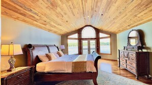 A bedroom with a wooden ceiling, a large bed, a dresser, and a view of a lake through a window. - Luxury Waterfront Home, Sleeps 14, 4BR + Bonus Room, Private Beach & Pickleball (Port Orchard)