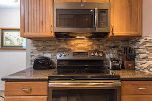 A kitchen with wooden cabinets, a stainless steel microwave, a stove, and a toaster. - Mahana Villas | Oceanfront 4 BR Sleeps 13 | Car Incl. w/ 6+ Nights | MAH ML-3728 by KBM (Lahaina)
