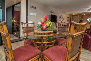 A round glass table surrounded by wicker chairs with red cushions, a floral centerpiece, and a television in the background. - Mahana Villas | Oceanfront 4 BR Sleeps 13 | Car Incl. w/ 6+ Nights | MAH ML-3728 by KBM (Lahaina)