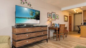 A living room with a flat-screen TV, wicker furniture, a dining table, chairs, and framed pictures on the wall. - Mahana Villas | Oceanfront 4 BR Sleeps 13 | Car Incl. w/ 6+ Nights | MAH ML-3728 by KBM (Lahaina)