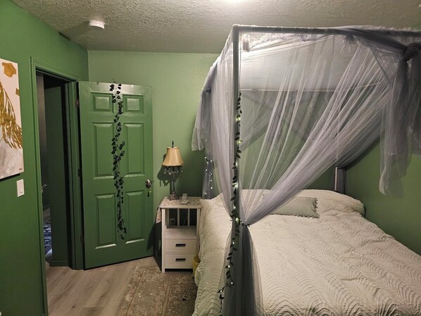A bedroom with a canopy bed, a green door with a plant design, a bedside table with a lamp, and a small nightstand.