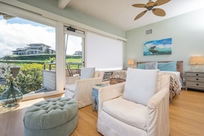 A bedroom with a bed, two chairs, a side table, a lamp, a ceiling fan, and a large window with a view of a building and greenery. - Kapalua Bay | Ocean View 6 BR, Sleeps 20 | Car Incl. w/ 6+ Nights | KBV ML-3734 by KBM (Lahaina)