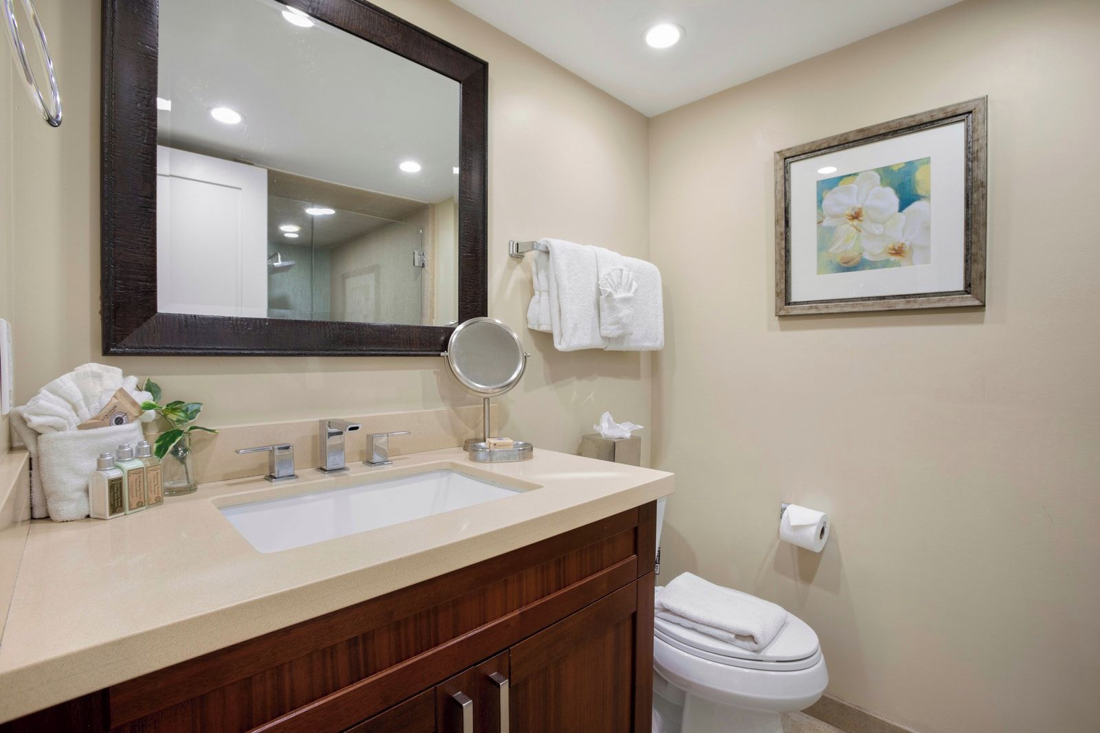 A bathroom with a large mirror, a sink with two faucets, a toilet, and a framed picture on the wall.