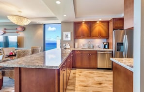A modern kitchen with wooden cabinets, a stainless steel refrigerator, and a large island with a granite countertop. - Mahana Villas | Oceanfront 4 BR Sleeps 12 | Car Incl. w/ 6+ Nights | MAH ML-3747 by KBM (Lahaina)