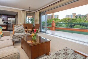 Ein modernes Wohnzimmer mit Sofa, Sessel und Couchtisch, einer Küchenzeile und einem Balkon mit Blick auf Gebäude und Grünflächen.