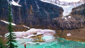 Gletsjer met een persoon die staat bij een turquoise meer, omringd door rotsachtig terrein en sneeuw.