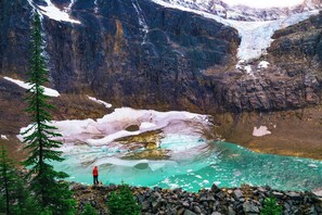 Ein Gletscher mit einer Person, die an einem türkisblauen See steht, umgeben von felsigem Gelände und Schnee.