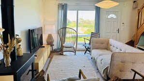 Sala de estar com sofá, cadeiras, televisão, abajur e vista por uma porta.