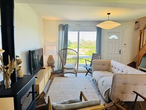 A living room with a sofa, chairs, a TV, a lamp, and a view through a door. - Maison de village 8 pers, au calme, jardin, wifi (QUIBERON)