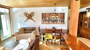 Un plafond en bois, une table à manger avec des chaises, un canapé et une œuvre d’art accrochée au mur.