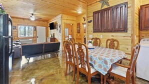 Ein Holzblockhaus-Interieur mit Esstisch, Stühlen, einer Bank, einem Kühlschrank und einem Deckenventilator.