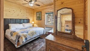 Camera da letto in una baita di legno con un letto, una cassettiera, uno specchio, una ventola a soffitto e una finestra.