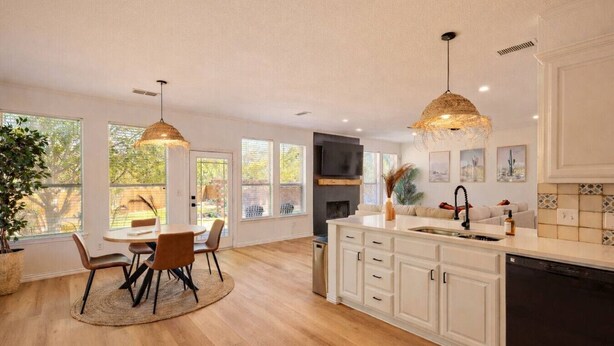 A modern kitchen with wooden flooring, white cabinetry, a central island with a sink, pendant lights, and a dining area with a round table and chairs.