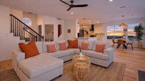 A modern living room with a sectional sofa, a dining area with a table and chairs, a kitchen in the background, and a view of the sunset through large windows.