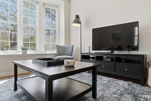 A modern living room with a dark wooden table, a black TV stand, and a large window with a view of trees. - Downtown Rochester Gem (Rochester)
