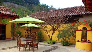Une terrasse avec une table et des chaises, un parasol vert et un mur jaune surmonté d’un toit en tuiles.