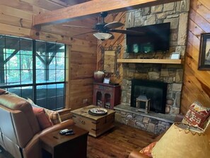 Ein Blockhaus-Innenraum mit Kamin aus Stein, einem darüber montierten Fernseher und einem großen Fenster.