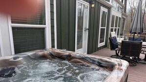Uma banheira de hidromassagem em um deck de madeira, com uma cadeira e um guarda-sol, cercada por uma casa com revestimento verde e detalhes brancos.