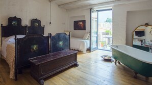 A bedroom with a wooden floor, a large bed with a headboard, a bench, a bathtub, and a balcony with outdoor furniture. - Authentic Sicilian house in village 20 km from the sea (Gratteri)