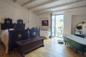 Une chambre avec un plancher en bois, un grand lit avec une tête de lit, un banc, une baignoire et un balcon agrémenté de mobilier d’extérieur.