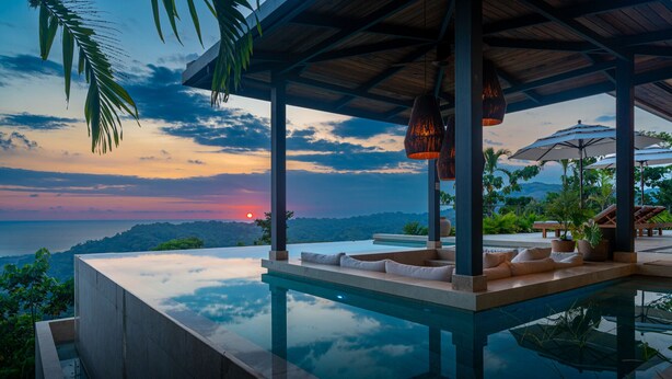 An infinity pool with a view of the sunset over the ocean, surrounded by lounge chairs and umbrellas. - Riverstone – Elevated Living Near Beach & Town (Dominical)