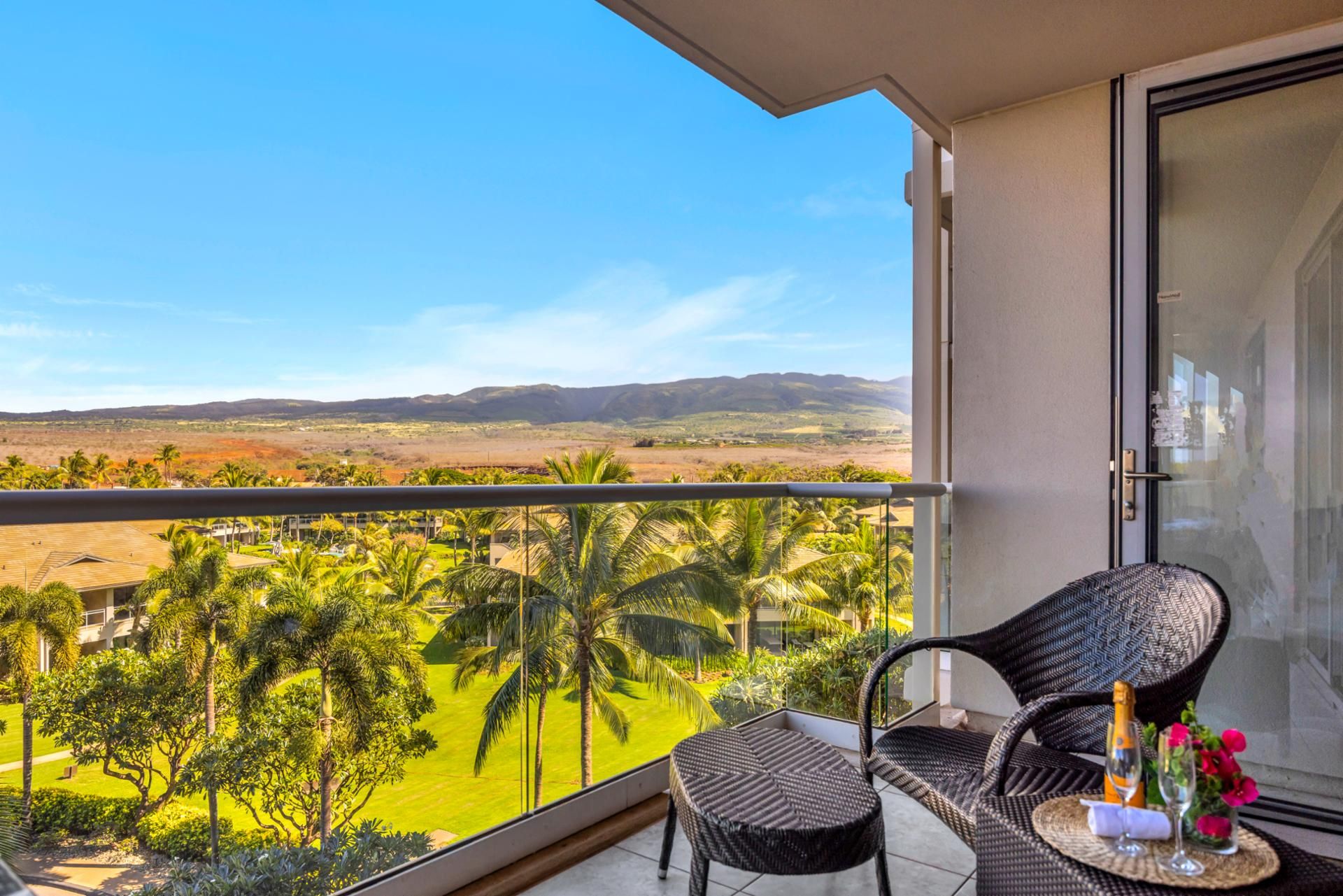 A balcony with a view of palm trees and distant hills, a wicker chair, and a table with a bottle and flowers.