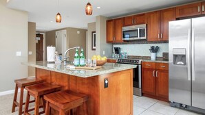 A kitchen with wooden cabinets, a stainless steel refrigerator, a microwave, and a granite countertop. - Honua Kai | Ocean View 3 BR, Sleeps 10 | Car Incl. w/ 6+ Nights | HKK ML-3902 by KBM (Lahaina)