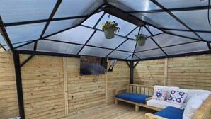 Un terrazzo in legno con tetto in vetro, due panche di legno con cuscini e vasi di fiori sospesi.